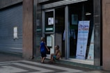 Two children are outside a building entrance with a sign indicating a kids' sale event scheduled for October 31, 2022. The building has a large, closed metal shutter, and the children are engaged in conversation or observation.