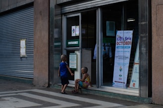 Two children are outside a building entrance with a sign indicating a kids' sale event scheduled for October 31, 2022. The building has a large, closed metal shutter, and the children are engaged in conversation or observation.