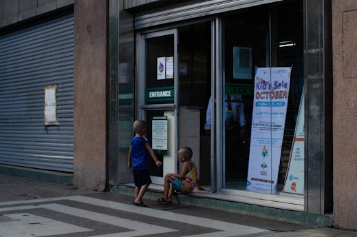 Two children are outside a building entrance with a sign indicating a kids' sale event scheduled for October 31, 2022. The building has a large, closed metal shutter, and the children are engaged in conversation or observation.