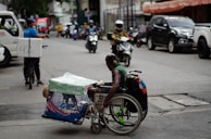 A man in a wheelchair navigates a busy urban street while carrying a pet dog in a cage attached to the wheelchair. The street is lined with parked cars and motorcycles, and other vehicles, such as a bicycle with a delivery box, are in motion. The environment appears bustling with activity, capturing a moment of urban life.