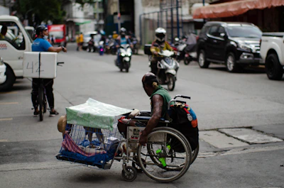 A man in a wheelchair navigates a busy urban street while carrying a pet dog in a cage attached to the wheelchair. The street is lined with parked cars and motorcycles, and other vehicles, such as a bicycle with a delivery box, are in motion. The environment appears bustling with activity, capturing a moment of urban life.