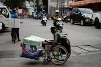 A man in a wheelchair navigates a busy urban street while carrying a pet dog in a cage attached to the wheelchair. The street is lined with parked cars and motorcycles, and other vehicles, such as a bicycle with a delivery box, are in motion. The environment appears bustling with activity, capturing a moment of urban life.