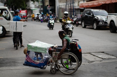 A man in a wheelchair navigates a busy urban street while carrying a pet dog in a cage attached to the wheelchair. The street is lined with parked cars and motorcycles, and other vehicles, such as a bicycle with a delivery box, are in motion. The environment appears bustling with activity, capturing a moment of urban life.