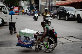 A man in a wheelchair navigates a busy urban street while carrying a pet dog in a cage attached to the wheelchair. The street is lined with parked cars and motorcycles, and other vehicles, such as a bicycle with a delivery box, are in motion. The environment appears bustling with activity, capturing a moment of urban life.