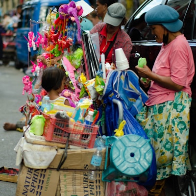 A street vendor setup with a variety of colorful toys and household items. On the left, a young child is looking at the display, while two women on the right appear to be managing the stall. Plastic cups and various packaged goods hang from the stall. The vivid mix of colors from the toys and items creates a vibrant and busy scene.