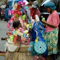 A street vendor setup with a variety of colorful toys and household items. On the left, a young child is looking at the display, while two women on the right appear to be managing the stall. Plastic cups and various packaged goods hang from the stall. The vivid mix of colors from the toys and items creates a vibrant and busy scene.