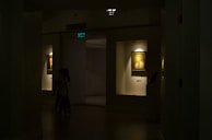 A dimly lit gallery interior features framed portraits on the walls, with soft light highlighting the artwork. Two paintings are displayed within recessed, illuminated niches. A pair of people is seen walking through the shadowed area of the gallery.