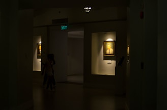 A dimly lit gallery interior features framed portraits on the walls, with soft light highlighting the artwork. Two paintings are displayed within recessed, illuminated niches. A pair of people is seen walking through the shadowed area of the gallery.