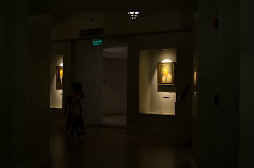 A dimly lit gallery interior features framed portraits on the walls, with soft light highlighting the artwork. Two paintings are displayed within recessed, illuminated niches. A pair of people is seen walking through the shadowed area of the gallery.