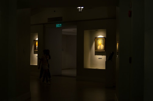 A dimly lit gallery interior features framed portraits on the walls, with soft light highlighting the artwork. Two paintings are displayed within recessed, illuminated niches. A pair of people is seen walking through the shadowed area of the gallery.