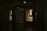 A dimly lit gallery interior features framed portraits on the walls, with soft light highlighting the artwork. Two paintings are displayed within recessed, illuminated niches. A pair of people is seen walking through the shadowed area of the gallery.