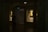 A dimly lit gallery interior features framed portraits on the walls, with soft light highlighting the artwork. Two paintings are displayed within recessed, illuminated niches. A pair of people is seen walking through the shadowed area of the gallery.