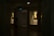 A dimly lit gallery interior features framed portraits on the walls, with soft light highlighting the artwork. Two paintings are displayed within recessed, illuminated niches. A pair of people is seen walking through the shadowed area of the gallery.