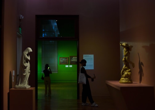 A dimly lit museum gallery featuring two sculptures; one large white sculpture on a pedestal and another golden sculpture casting a shadow on the wall. A person stands to the left, looking at their phone, while another person on the right is holding an object against a backdrop of a green-painted room with framed artwork.