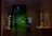 A dimly lit museum gallery featuring two sculptures; one large white sculpture on a pedestal and another golden sculpture casting a shadow on the wall. A person stands to the left, looking at their phone, while another person on the right is holding an object against a backdrop of a green-painted room with framed artwork.