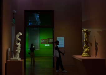 A dimly lit museum gallery featuring two sculptures; one large white sculpture on a pedestal and another golden sculpture casting a shadow on the wall. A person stands to the left, looking at their phone, while another person on the right is holding an object against a backdrop of a green-painted room with framed artwork.