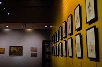 A gallery wall featuring a series of framed black-and-white sketches is displayed under soft lighting. The wall is painted a vibrant yellow, adding a contrasting backdrop to the monochromatic art pieces. Another section of the wall, painted in a neutral tone, exhibits framed paintings and plaques, creating a dedicated exhibition space.