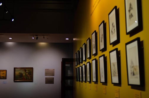 A gallery wall featuring a series of framed black-and-white sketches is displayed under soft lighting. The wall is painted a vibrant yellow, adding a contrasting backdrop to the monochromatic art pieces. Another section of the wall, painted in a neutral tone, exhibits framed paintings and plaques, creating a dedicated exhibition space.