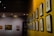 A gallery wall featuring a series of framed black-and-white sketches is displayed under soft lighting. The wall is painted a vibrant yellow, adding a contrasting backdrop to the monochromatic art pieces. Another section of the wall, painted in a neutral tone, exhibits framed paintings and plaques, creating a dedicated exhibition space.