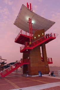 A modern architectural structure featuring a large white canopy supported by red metal beams. The structure includes multiple levels accessible by red stairs, with a few people standing on the balconies. The setting appears to be by a waterfront, surrounded by a vibrant evening sky with shades of pink.