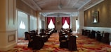 A spacious and elegant dining area with rows of tables and chairs covered in dark cloth. The room is well-lit with natural light coming through large windows with red curtains. The ceiling is intricately designed with chandeliers, and the floor is covered in a colorful floral-patterned carpet. The walls are adorned with framed artwork, and there is a luxurious ambiance throughout the space.