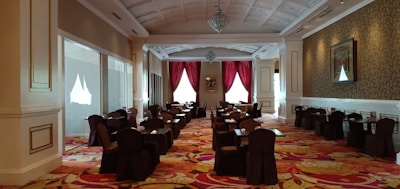 A spacious and elegant dining area with rows of tables and chairs covered in dark cloth. The room is well-lit with natural light coming through large windows with red curtains. The ceiling is intricately designed with chandeliers, and the floor is covered in a colorful floral-patterned carpet. The walls are adorned with framed artwork, and there is a luxurious ambiance throughout the space.
