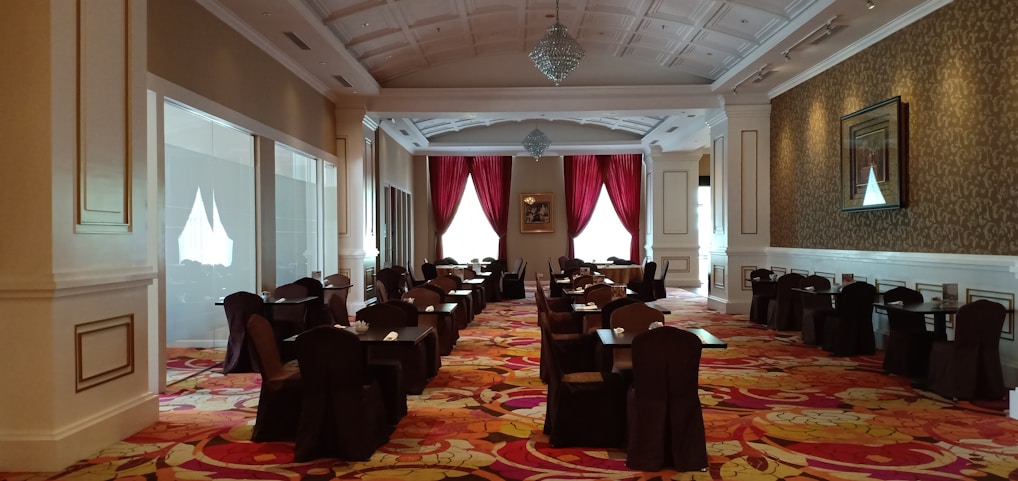 A spacious and elegant dining area with rows of tables and chairs covered in dark cloth. The room is well-lit with natural light coming through large windows with red curtains. The ceiling is intricately designed with chandeliers, and the floor is covered in a colorful floral-patterned carpet. The walls are adorned with framed artwork, and there is a luxurious ambiance throughout the space.