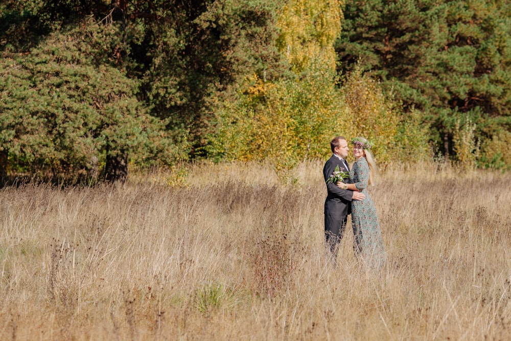 a man and woman standing in a field of tall grass with trees in the background