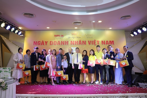 A group of people dressed in semi-formal attire standing on a stage, each holding a gift bag with a circular logo. The background features a yellow banner with red text and several logos. Stage lights are positioned above, and confetti is scattered on the floor.