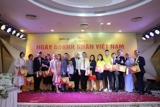 A group of people dressed in semi-formal attire standing on a stage, each holding a gift bag with a circular logo. The background features a yellow banner with red text and several logos. Stage lights are positioned above, and confetti is scattered on the floor.