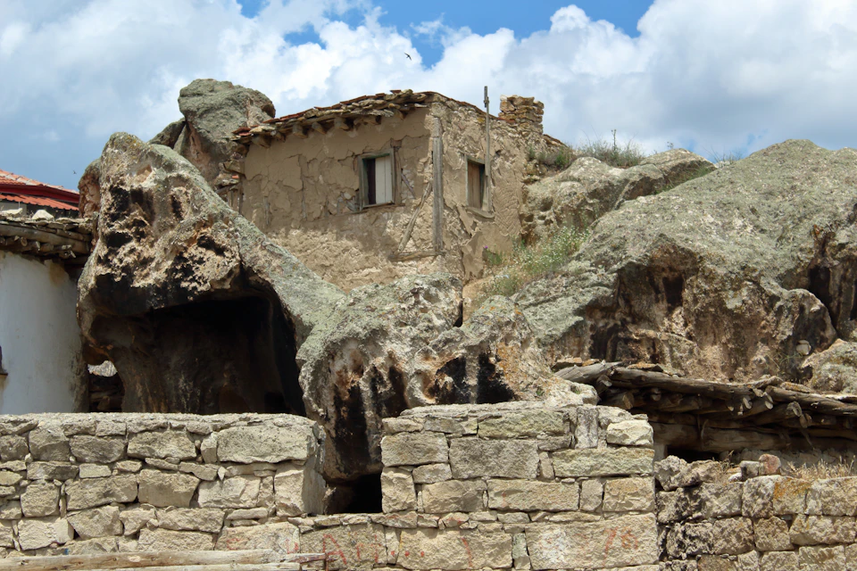 The image depicts a small, rustic stone house nestled on a rocky cliff face. The house features a thatched roof and is surrounded by an array of rocks and boulders, creating a rugged and natural setting. A stone wall can be seen in the foreground, adding to the overall sense of the scene. The sky above is filled with clouds, suggesting a cloudy day or overcast weather conditions.