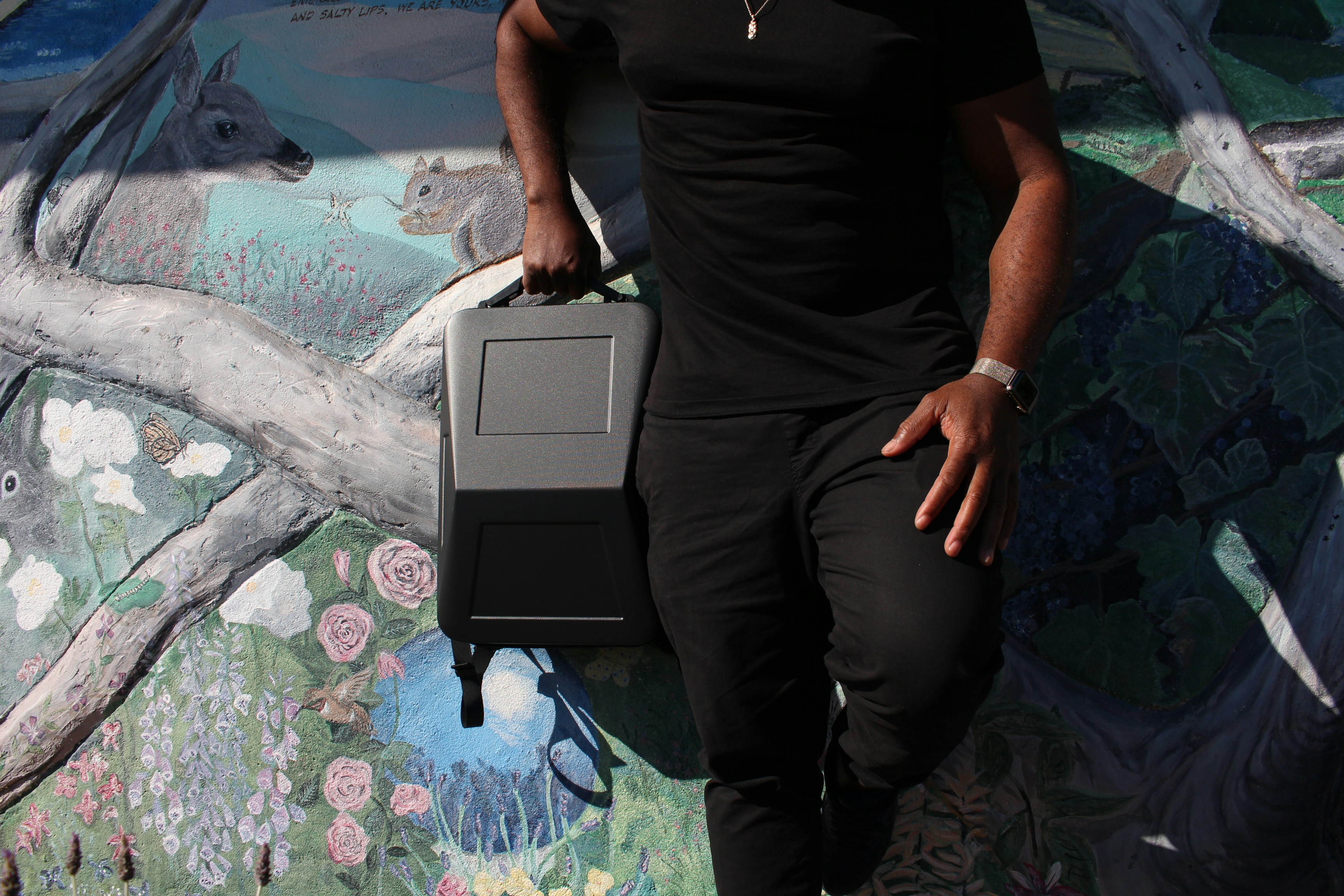 a person holding a box, Model leaning against a mural while carrying a Cyberbackpack Tesla Cybertruck anti-theft laptop travel backpack bag by Cyberbackpack.com. The Cyberbackpack is a modern anti-theft hardshell slim laptop backpack that is perfect for travel, work, and play, with enough space to carry up to a 17-inch laptop, books, and anything that you may need for your busy day. This modern slim backpack is lightweight, weighing less than 2.9 lbs, and is ergonomically designed to protect your back and shoulders. special features include a USB charging port, powerbank, and TSA-approved passcode lock.