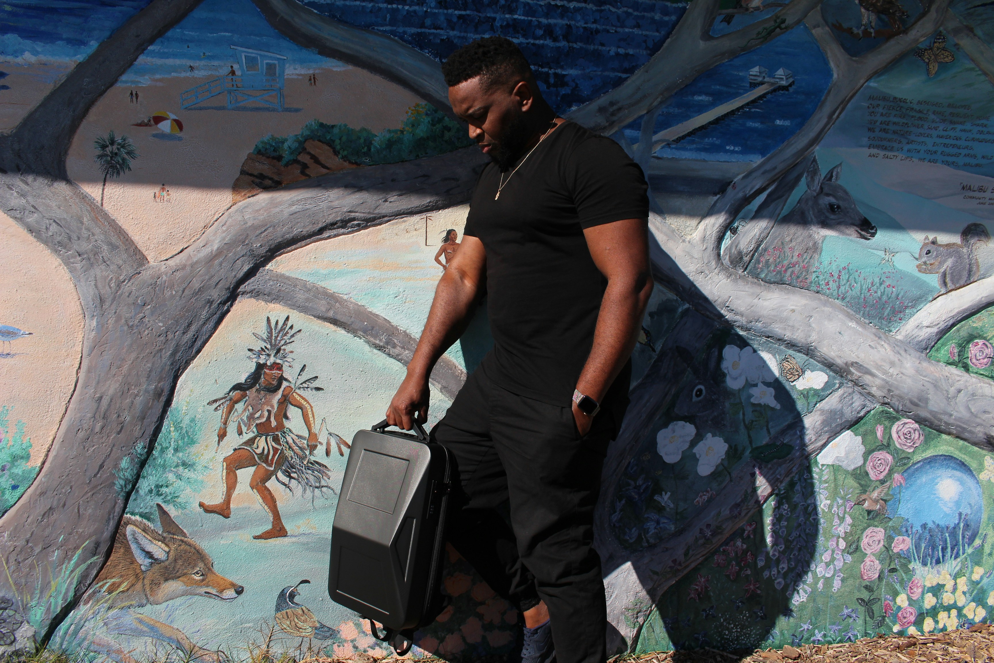 a man holding a suitcase, Black guy by a Malibu beach ocean mural while carrying a Cyberbackpack Tesla Cybertruck anti-theft laptop travel backpack bag by Cyberbackpack.com. The Cyberbackpack is a modern anti-theft hardshell slim laptop backpack that is perfect for travel, work, and play, with enough space to carry up to a 17-inch laptop, books, and anything that you may need for your busy day. This modern slim backpack is lightweight, weighing less than 2.9 lbs, and is ergonomically designed to protect your back and shoulders. special features include a USB charging port, powerbank, and TSA-approved passcode lock.