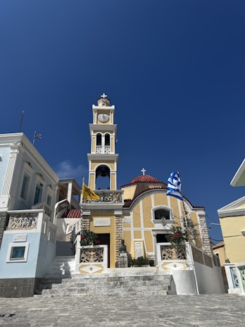 A picturesque church with a prominent clock tower and red-tiled roof set against a clear blue sky. The church features white and yellow walls, ornate railings, and a set of stone steps leading up to the entrance. A Greek flag and another flag are displayed on poles near the building.
