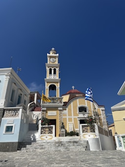 A picturesque church with a prominent clock tower and red-tiled roof set against a clear blue sky. The church features white and yellow walls, ornate railings, and a set of stone steps leading up to the entrance. A Greek flag and another flag are displayed on poles near the building.