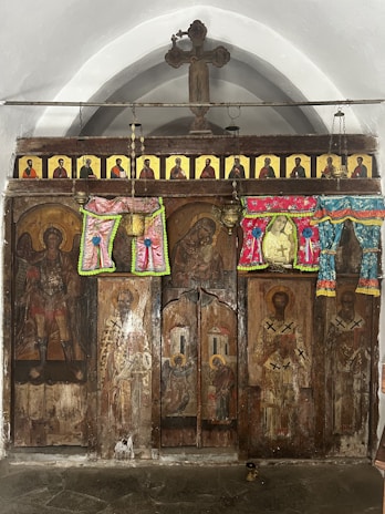 A religious scene with a wooden panel decorated with painted icons of saints and other religious figures. There are colorful fabric decorations and hanging lamps, and a large cross at the top. The panel appears weathered, suggesting age.