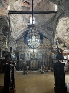 A richly decorated interior of a historic church with ornate religious iconography covering the walls. The ceiling features intricate stonework and a large chandelier hanging in the center. There are dark wooden accents and stands, with small crosses placed on top. Various candelabras and religious artifacts are placed around the area.