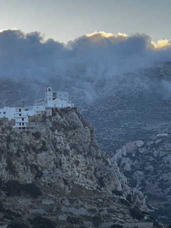 A white, multi-level church perched on the edge of a steep, rocky mountain with a dramatic and cloud-filled sky in the background. The rugged landscape is dotted with patches of greenery.