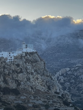 A white, multi-level church perched on the edge of a steep, rocky mountain with a dramatic and cloud-filled sky in the background. The rugged landscape is dotted with patches of greenery.