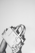 A person holds a stylish Prada handbag with both hands. The handbag is elegantly designed and features a prominent logo. The image is in black and white, adding a classic and sophisticated feel. The background is plain, emphasizing the focus on the handbag.