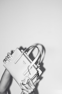 A person holds a stylish Prada handbag with both hands. The handbag is elegantly designed and features a prominent logo. The image is in black and white, adding a classic and sophisticated feel. The background is plain, emphasizing the focus on the handbag.