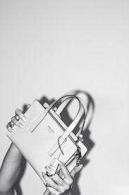 A person holds a stylish Prada handbag with both hands. The handbag is elegantly designed and features a prominent logo. The image is in black and white, adding a classic and sophisticated feel. The background is plain, emphasizing the focus on the handbag.