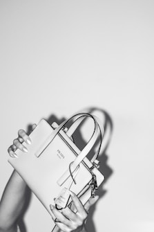 A person holds a stylish Prada handbag with both hands. The handbag is elegantly designed and features a prominent logo. The image is in black and white, adding a classic and sophisticated feel. The background is plain, emphasizing the focus on the handbag.