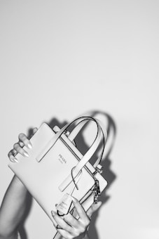 A person holds a stylish Prada handbag with both hands. The handbag is elegantly designed and features a prominent logo. The image is in black and white, adding a classic and sophisticated feel. The background is plain, emphasizing the focus on the handbag.