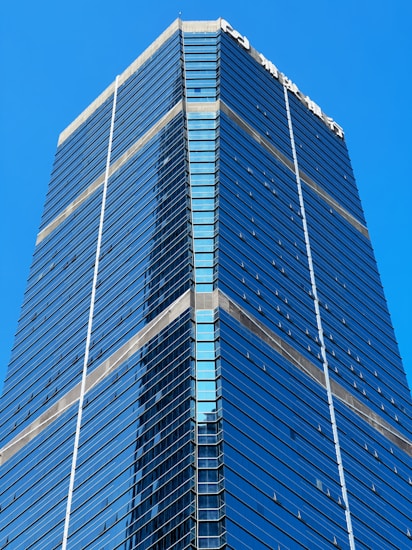 A tall, modern skyscraper with a sleek design featuring glass and steel elements. The building's facade is composed of reflective blue-tinted windows that stretch upwards to the sky. The structure has sharp angles and a symmetrical appearance, and on top, there is white text, possibly indicating a company name.