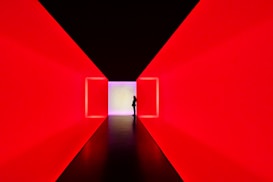 A dark silhouetted person stands in a strikingly lit corridor with walls glowing in intense red, leading to a distant square aperture emitting a soft white light.