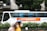 A bus with the logo and branding of FPT University is parked on the side of the street. The bus is white with large windows, blue curtains, and a prominent orange banner along the side. The background features a modern building and lush green trees, with some blurred motion in the foreground likely due to moving people.