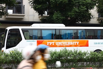 A bus with the logo and branding of FPT University is parked on the side of the street. The bus is white with large windows, blue curtains, and a prominent orange banner along the side. The background features a modern building and lush green trees, with some blurred motion in the foreground likely due to moving people.