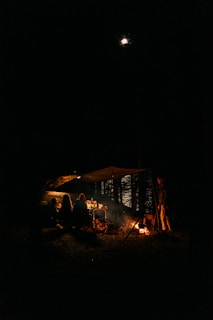 A nighttime camping scene with three people sitting under a canopy next to a tent and a car. They are gathered around a small campfire, with a table visible behind them holding various items. One person is standing near the fire. The moon is shining in the night sky above, and trees are faintly visible in the background.
