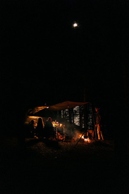 A nighttime camping scene with three people sitting under a canopy next to a tent and a car. They are gathered around a small campfire, with a table visible behind them holding various items. One person is standing near the fire. The moon is shining in the night sky above, and trees are faintly visible in the background.